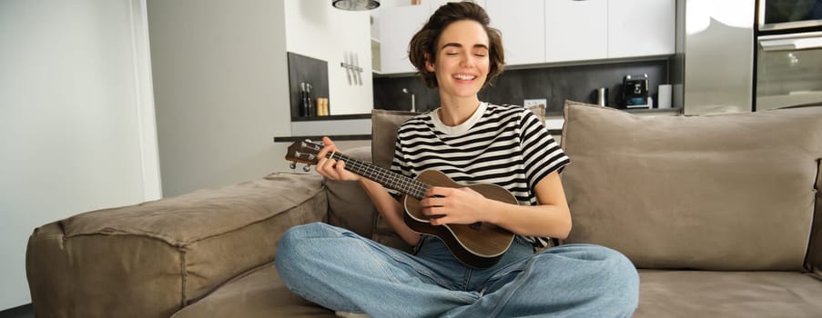 Portrait of cheerful young woman playing her ukulele, singing and laughing, sitting in living room at home. Lifestyle and music concept