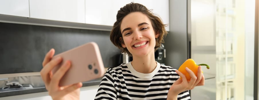 Close up portrait of cute brunette woman, taking selfie with yellow fresh pepper, recording video on smartphone, posing with vegetables in the kitchen, making cooking blog on social media app.