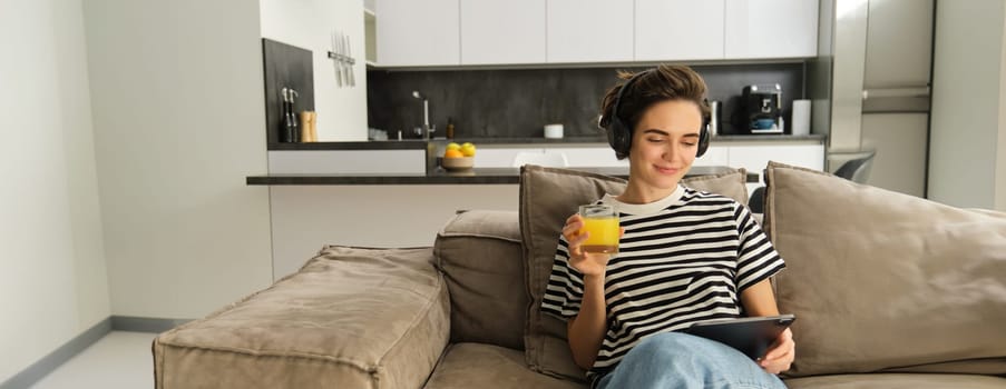 Portrait of woman in headphones, sitting on sofa with tablet and drinking orange juice, watching tv show on her gadget, using social media application, spending time at home.