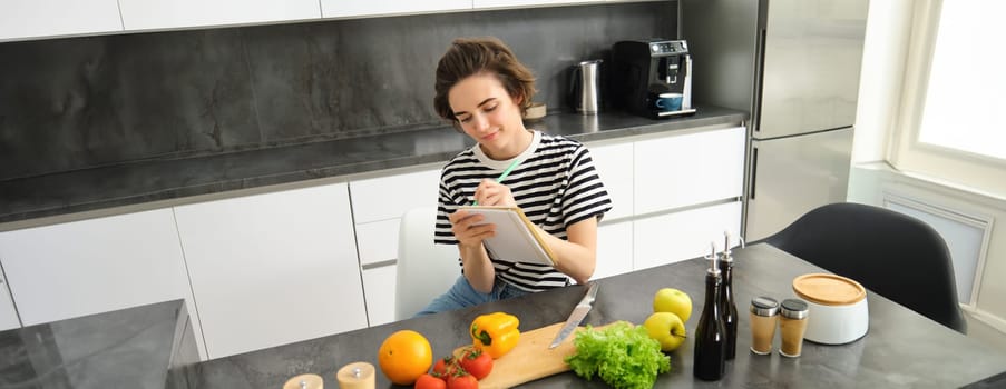 Portrait of woman writing down her recipe, making notes in notebook during cooking, sitting in the kitchen with chopping board and vegetables, preparing a meal.