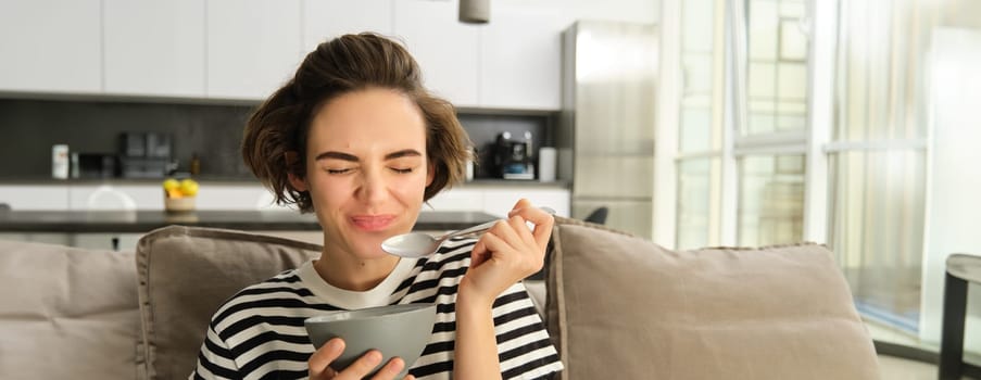 Portrait of young female model, student eating quick breakfast, holding bowl of granola or cereals with milk, sitting on sofa in living room.