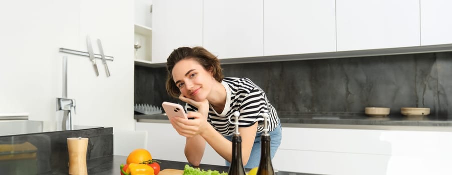 Portrait of woman, lifestyle blogger, posting picture of her cooking process on smartphone app, standing near vegetables and chopping board.