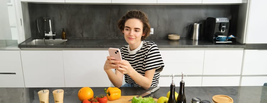 Portrait of young woman with smartphone, sitting in the kitchen and cooking salad, searching for healthy recipe online, has vegetables and chopping board on the table.