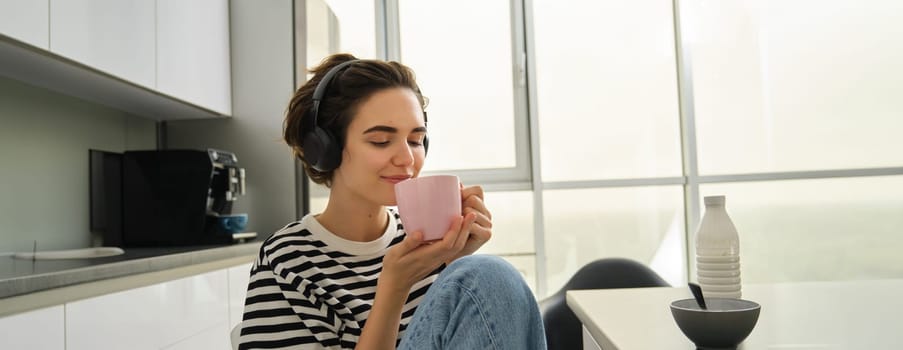 Portrait of tender, beautiful woman with tea, listens music in headphones, holds cup and smiles.