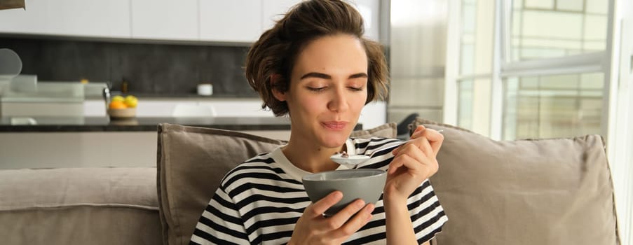 Portrait of young brunette woman eating on sofa, holding bowl and spoon, having cereals with milk for breakfast, enjoying her granola, spending time living room.