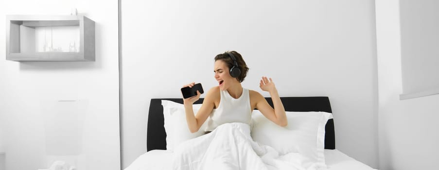 Carefree woman in bed, wakes up and listens to music in wireless headphones, dancing and singing, using mobile phone as microphone.