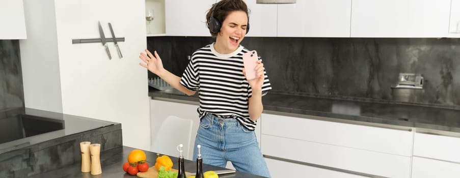 Young woman cooking meal in kitchen, listening to music in wireless headphones, singing into smartphone microphone, making salad, chopping vegetables.