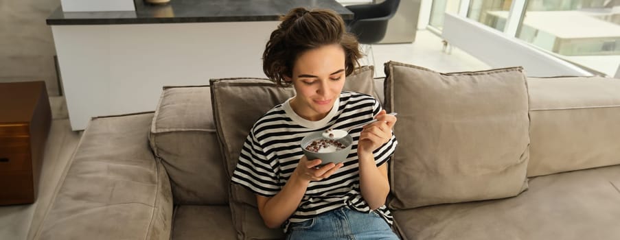 Portrait of young female model, student eating quick breakfast, holding bowl of granola or cereals with milk, sitting on sofa in living room.