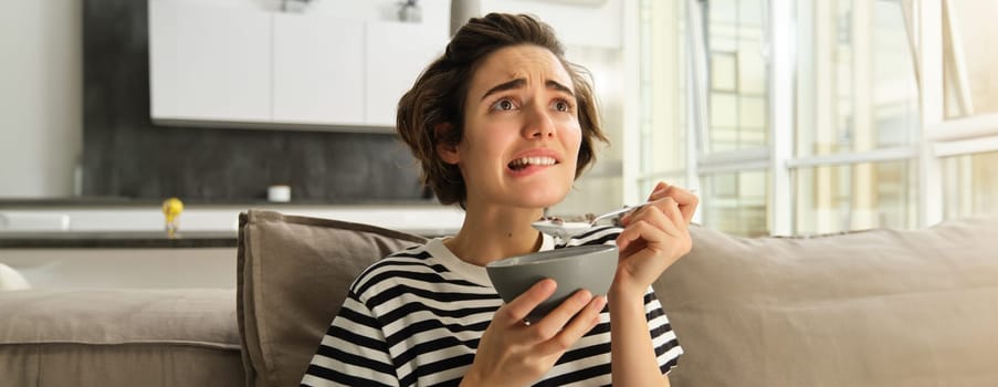 Close up portrait of female model, girl eating breakfast and watching intense scene in movie, staring at tv screen in living room, holding bowl of cereals with milk.