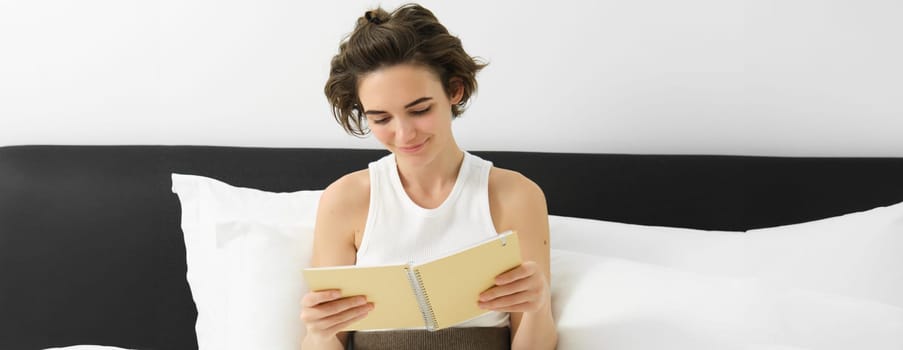 Portrait of woman reading her planner and starting the day, looking at notes, sitting on bed in morning.