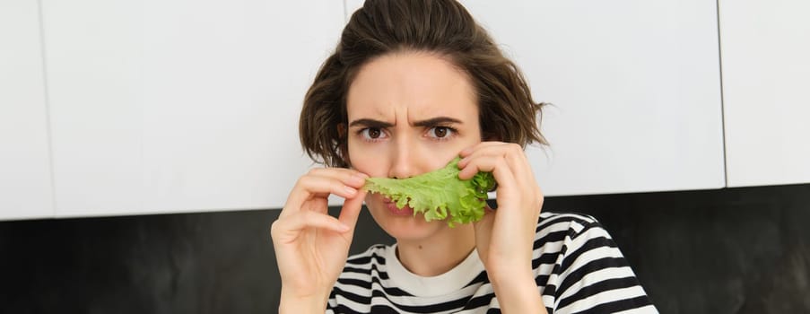 Frowning silly woman, holding lettuce leaf near face and frowning at camera, making pouting face, dislike eating vegetables.