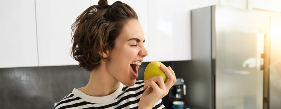 Close up portrait of smiling woman in the kitchen, holding an apple, eating fruit, having a healthy snack for lunch at home.