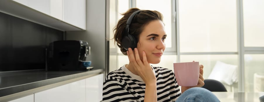 Close up portrait of smiling brunette woman, student drinks her tea and listens music or ebook in headphones, sits in kitchen and relaxes.