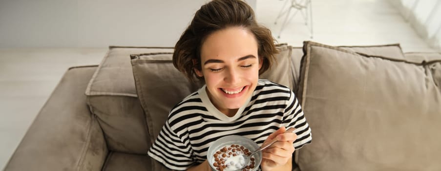Upper angle shot of happy, cute young woman on sofa, eating bowl of cereals with milk and smiling, enjoying her breakfast.
