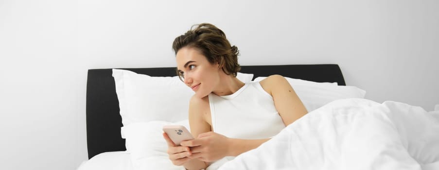 Portrait of young woman waking up in bed, using smartphone, turn off alarm on mobile phone, lying in her bedroom on white linen sheets.