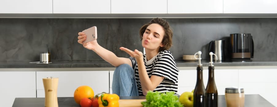 Cute young woman, lifestyle blogger cooking, sitting in the kitchen with vegetables and chopping board, taking selfie on her new smartphone, sending kisses at mobile camera.