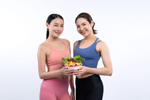 Two young sporty Asian women in sportswear holding salad bowl fill with fruit and vegetable. Natural youthful and fit body lifestyle people with balance nutrition on isolated background. Vigorous