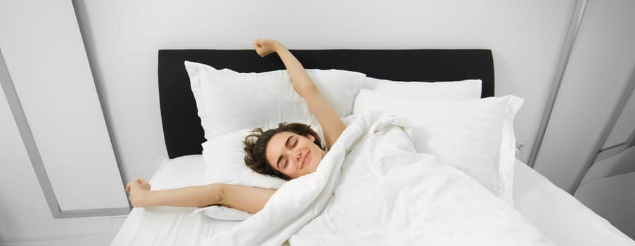 Portrait of happy, pleased young woman, stretching her arms with satisfied smile, lying in bed of soft pillow, waking up alone in bed, had good night sleep.