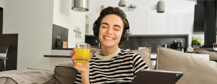 Close up portrait of smiling, happy brunette woman in living room, sitting on sofa and drinking orange juice, reading on digital tablet, using her gadget and resting at home.