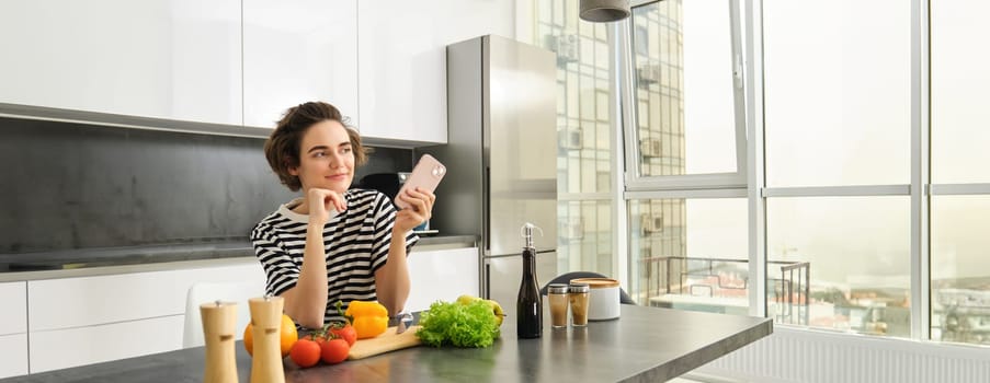 Portrait of young woman standing in the kitchen with vegetables and chopping board, holding smartphone, searching healthy diet recipes on mobile phone browser, cooking at home.