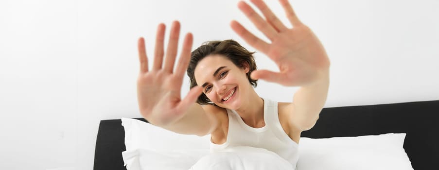 Portrait of sleepy, happy young woman, wakes up in her bed, stretching out hands, hiding face from camera, smiling and laughing.