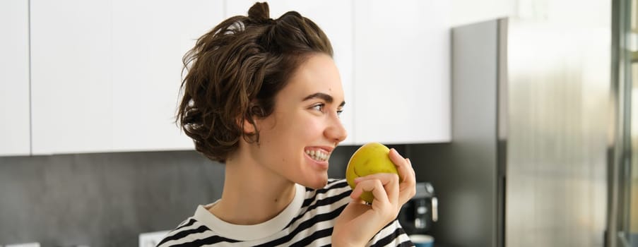 Close up portrait of cute young modern woman, eating green apple and looking aside, smiling while biting a fruit, having a snack in the kitchen.