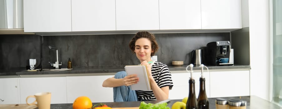 Portrait of young smiling woman cooking with vegetables, writing down ingredients, looking at her notebook, reading recipe, sitting in kitchen.