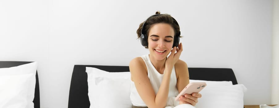 Portrait of smiling young woman in wireless headphones, sitting on bed with mobile phone, listening music in earphones.