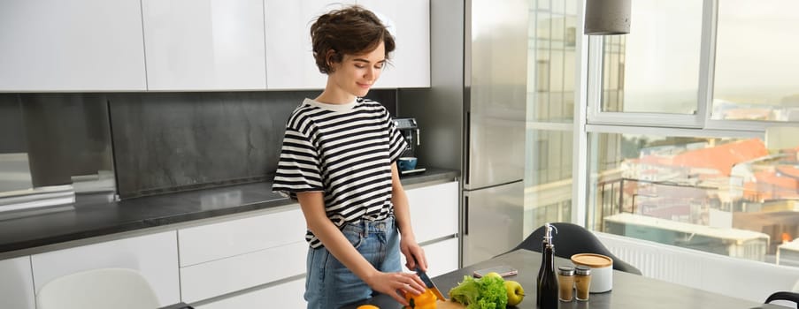 Portrait of smiling cute young woman making breakfast, chopping vegetables in the kitchen, preparing vegan meal.