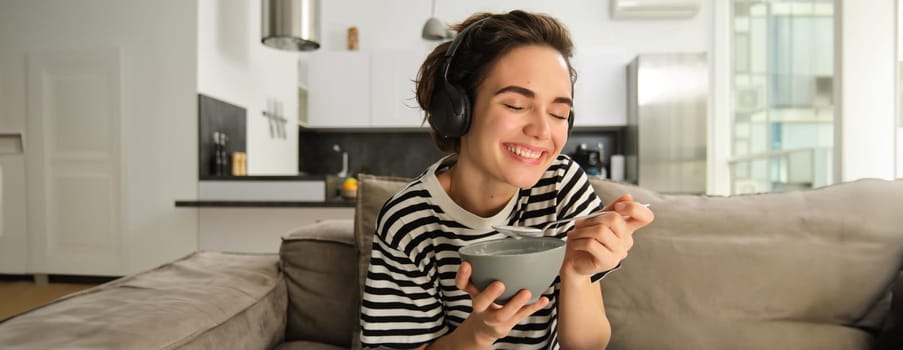 Close up of happy beautiful woman, laughing and smiling, watching tv in wireless headphones, holding bowl of cereals, having breakfast in living room on sofa.