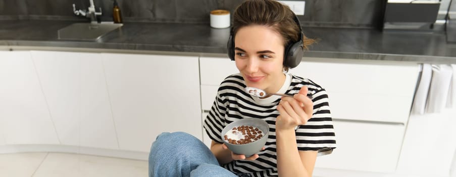 Young woman eating cereals in the morning, student having her breakfast, listening music or podcast in wireless headphones.