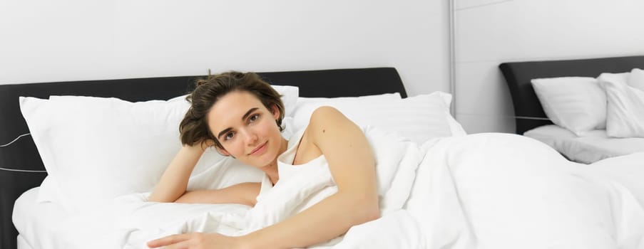 Portrait of beautiful woman in bed, waking up in morning, relaxing under white blanket with comfortable and soft pillows, looking happy and pleased.