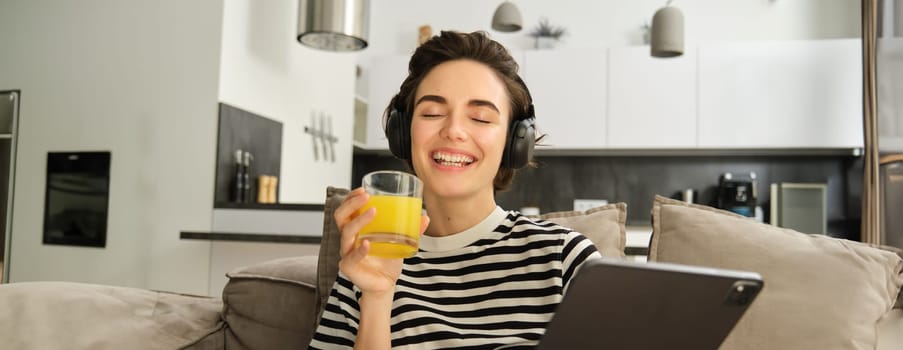 Close up portrait of young brunette woman in living room, watching funny video or tv series on digital tablet, drinking orange juice, sitting on sofa and laughing.
