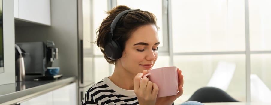 Portrait of smiling, relaxed woman, drinks warm tea and listens music or e-book in kitchen. Lifestyle and people concept