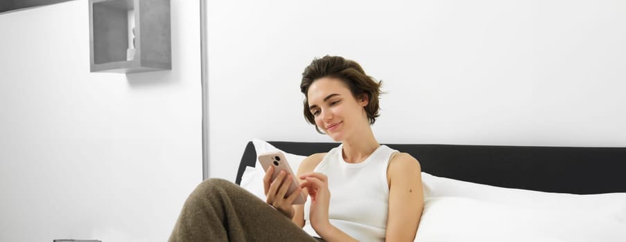 Portrait of young beautiful woman lying in bed, resting in bedroom, messaging, using mobile phone, holding smartphone and smiling, scrolling social media, spending comfort time at home.