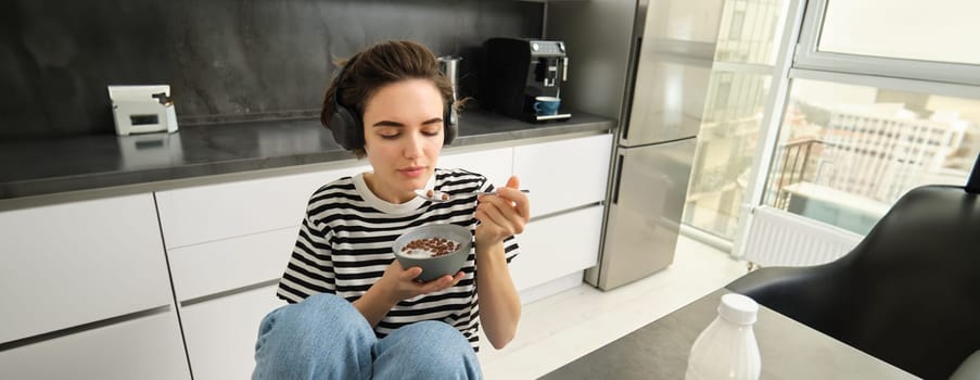 Happy brunette woman in the kitchen, eating cereals with milk for breakfast, listening music in headphones, having her morning meal.