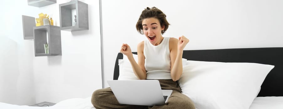 Portrait of woman sitting on bed, looking at laptop with excited, amazed face, celebrating, making fist pump, triumphing after winning on computer.