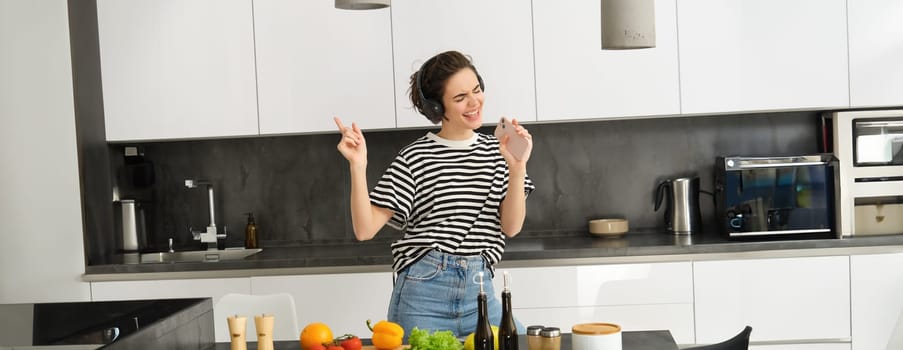 Portrait of happy girl cooking salad and listening to music in headphones, dancing while making a meal in the kitchen.