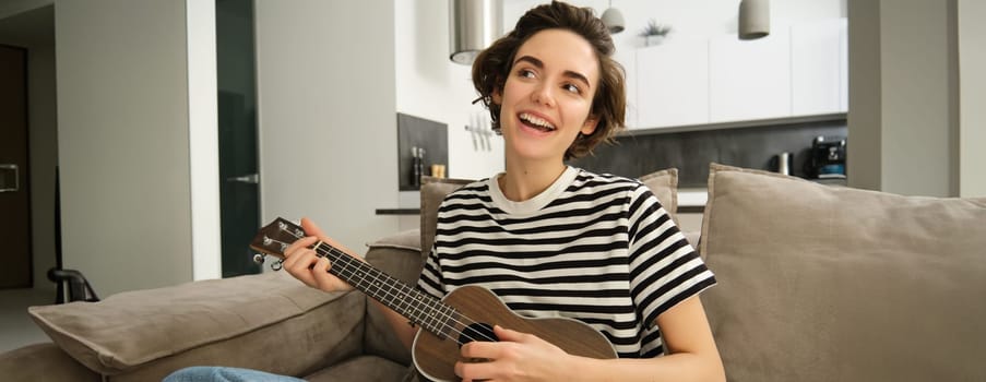 Beautiful young woman playing ukulele, singing and enjoying her musical instrument, spending time alone at home, relaxing in living room.