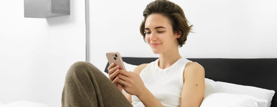 Cute young woman with mobile phone in hands, lying in bed in tank top, looking at smartphone with pleased smile.