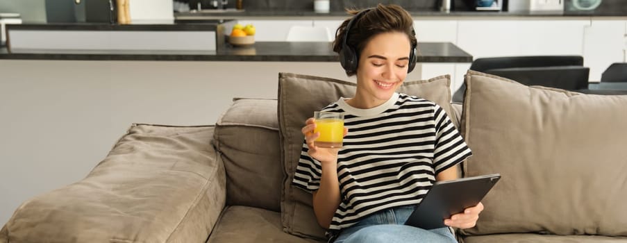 Portrait of woman in headphones, sitting on sofa with tablet and drinking orange juice, watching tv show on her gadget, using social media application, spending time at home.