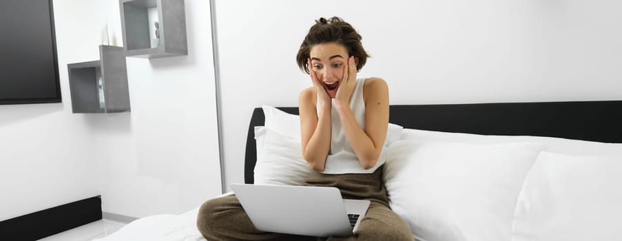 Image of woman at home, sitting on bed and staring shocked at laptop, looking at screen with surprised, happy face expression.