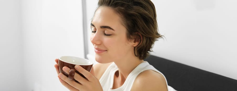 Sensual morning concept. Young beautiful woman drinking coffee in bed, holding mug and smelling nice aroma of freshly brewed drink.