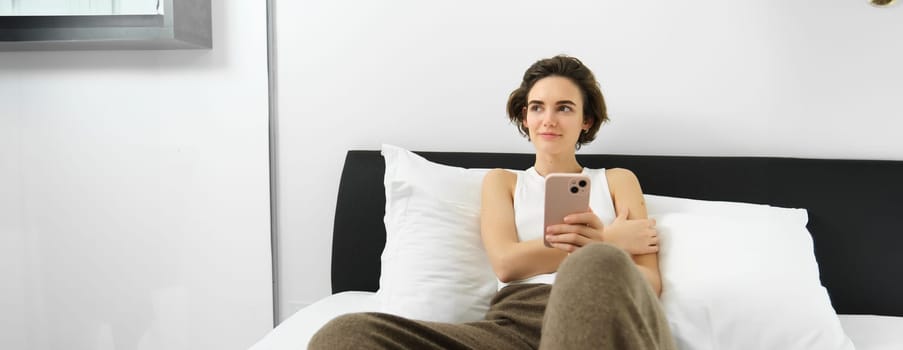 Lifestyle and technology concept. Smiling beautiful woman lying in bed with smartphone, resting in bedroom, chatting on social media, online shopping.