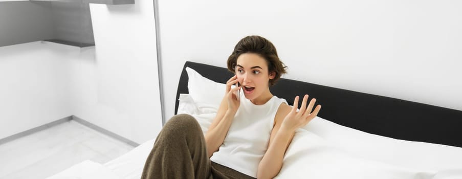 Chatty young woman laughing, talking on phone, calling friend, lying in bed and having conversation with someone, resting in bedroom.