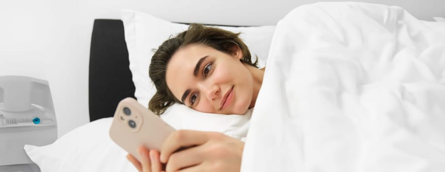 Smiling beautiful woman, lying in bed with smartphone, looking at mobile phone, using application, reading message and looking pleased.