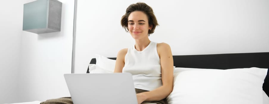 Portrait of smiling modern woman, sitting on her bed with laptop, looking happy, working from home, checking her emails on computer, browsing online shops.