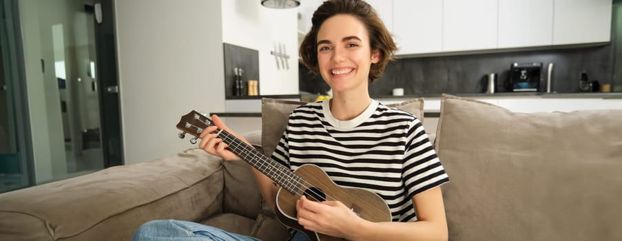 Beautiful girl playing ukulele on sofa, singing her favourite song and picking chords on small guitar, sitting in living room.