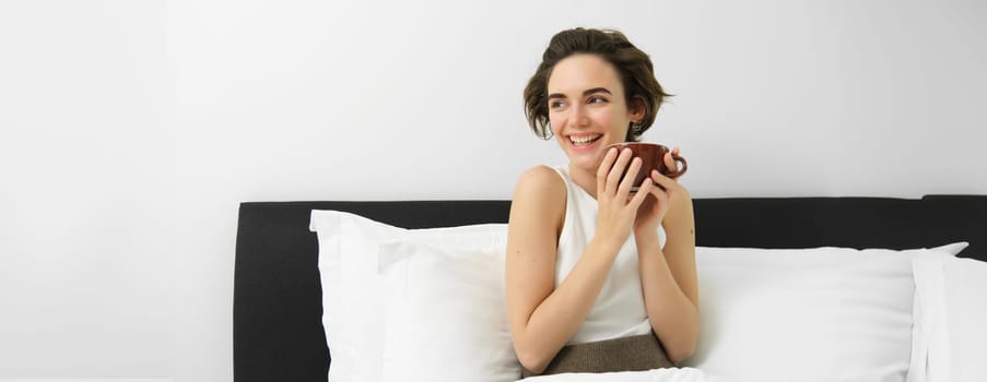 Portrait of happy, beautiful woman in bed, having her morning cup of coffee, drinking from mug and smiling, waking up and feeling joy.