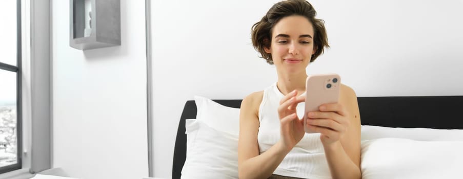 Portrait of smiling beautiful woman, sitting on bed with laptop, using smartphone, checking message on mobile phone, relaxing at home.